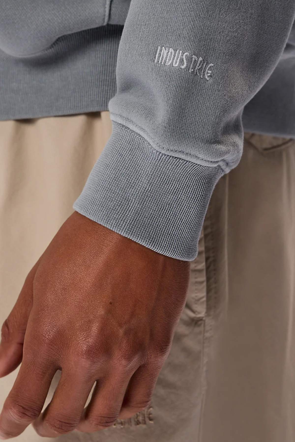 A close-up of a person’s hand and lower arm wearing the Industrie The Del Sur Washed Hoodie in gray with “INDUSTRIE” embroidered on the sleeve, paired with light beige shorts. The background is blurred.