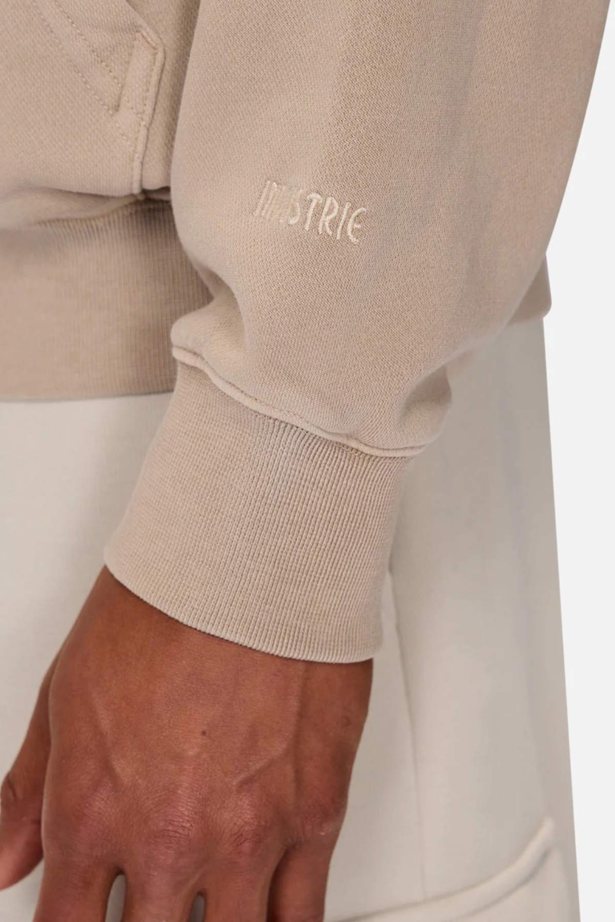 A close-up features someone in an oversized beige Industrie The Del Sur Washed Hoodie with "INDSTRIE" on the sleeve, paired with light-colored pants for a relaxed and effortlessly stylish look.