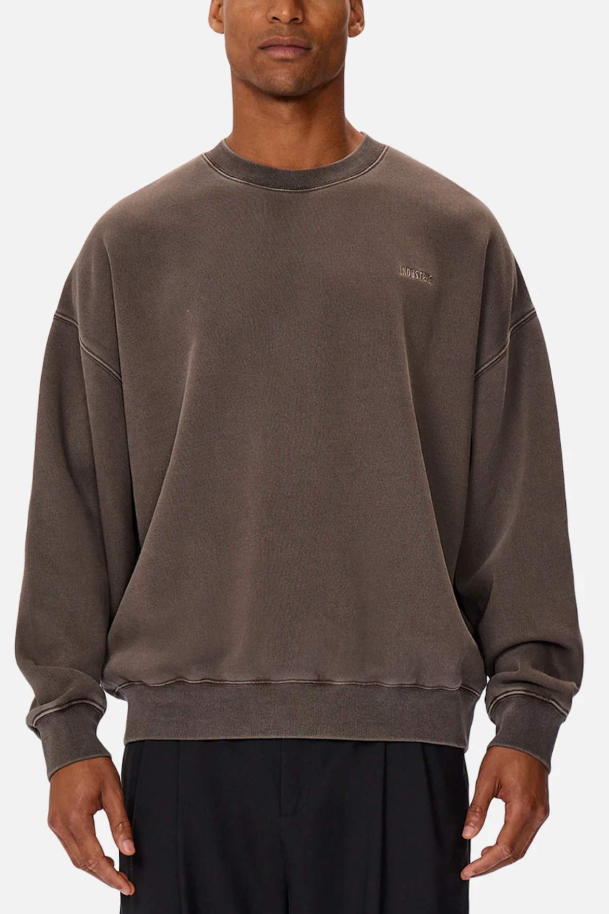 A man wears the Industrie The Del Sur Washed Sweat, an oversized brown crewneck jumper with subtle stitching and a discreet chest logo, paired with black pants against a plain white background.