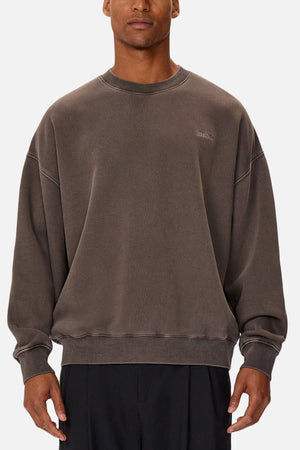 A man wears the Industrie The Del Sur Washed Sweat, an oversized brown crewneck jumper with subtle stitching and a discreet chest logo, paired with black pants against a plain white background.