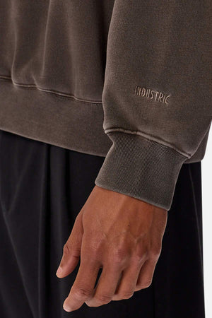 A close-up of a person’s hand and forearm wearing the Industrie The Del Sur Washed Sweat, a brown oversized jumper with "INDUSTRIE" embroidered on the cuff, paired with black pants against a plain light background.