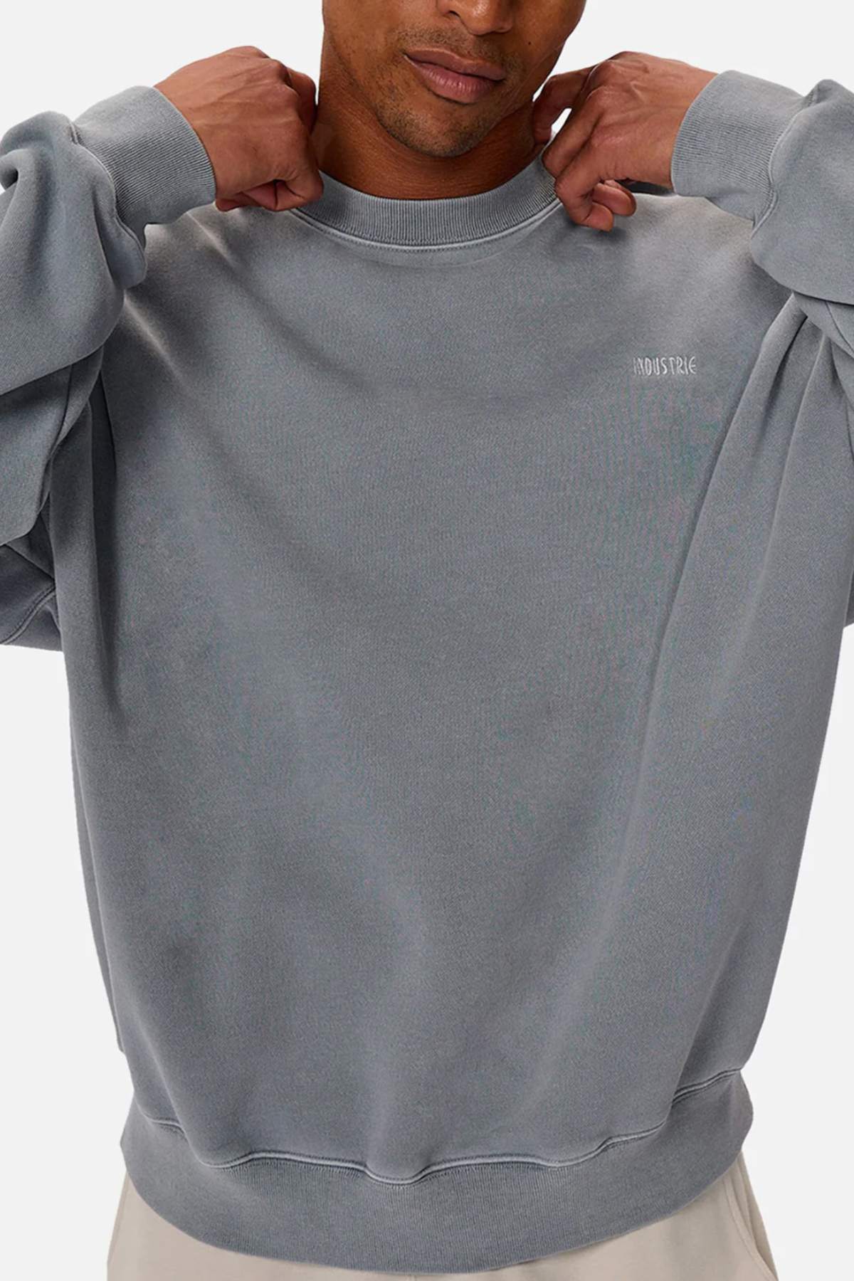 A person adjusts the collar of the Industrie The Del Sur Washed Sweat, an oversized grey jumper with long sleeves, rib knit cuffs, and small white chest text. Their face is partly out of frame.