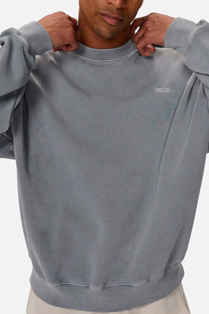 A person adjusts the collar of the Industrie The Del Sur Washed Sweat, an oversized grey jumper with long sleeves, rib knit cuffs, and small white chest text. Their face is partly out of frame.