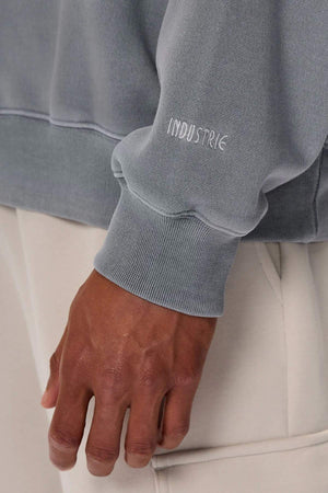A close-up shows a person’s hand and wrist in the Industrie The Del Sur Washed Sweat, a grey sweatshirt with "INDUSTRIE" embroidered on the rib knit cuff, paired with light beige pants.