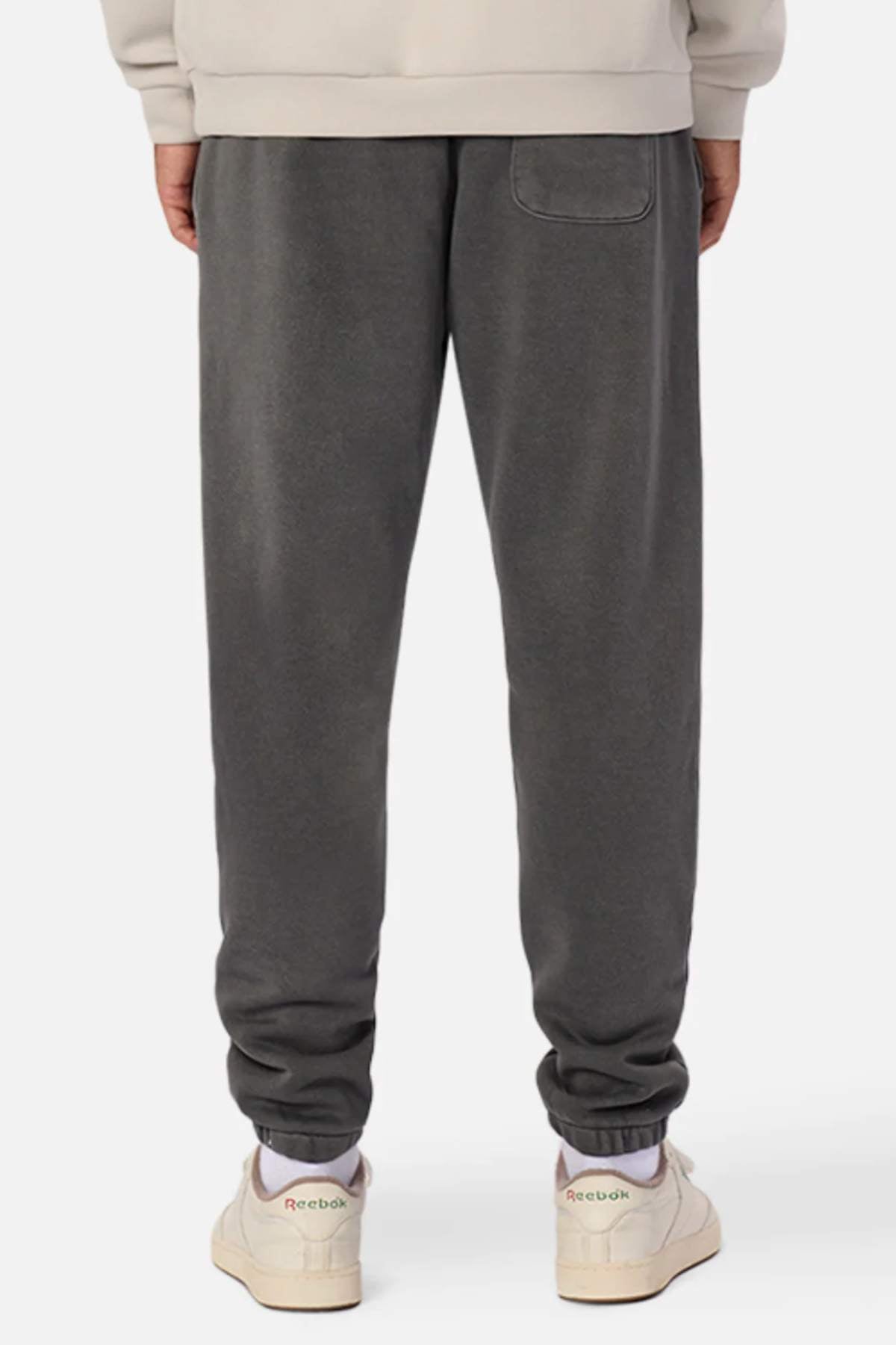 Rear view of a person wearing dark gray Industrie The Del Sur Washed Track Pant with an elastic drawstring waist and single back pocket, paired with a beige sweatshirt and off-white Reebok sneakers, standing against a plain white background.
