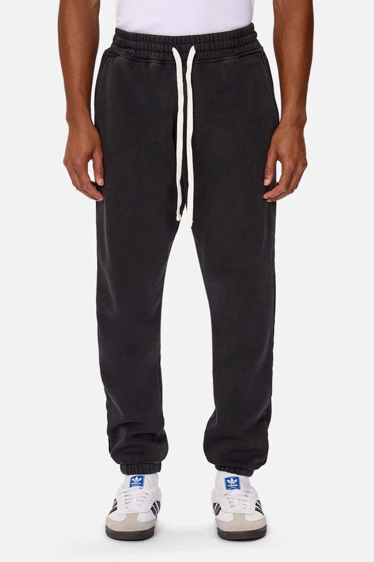 Someone is wearing black joggers with an elasticated drawstring waist and a white shirt, paired with white Adidas sneakers with blue stripes. These Industrie The Del Sur Washed Track Pants feature tapered ankles, adding a casual, comfortable touch against the plain white background.