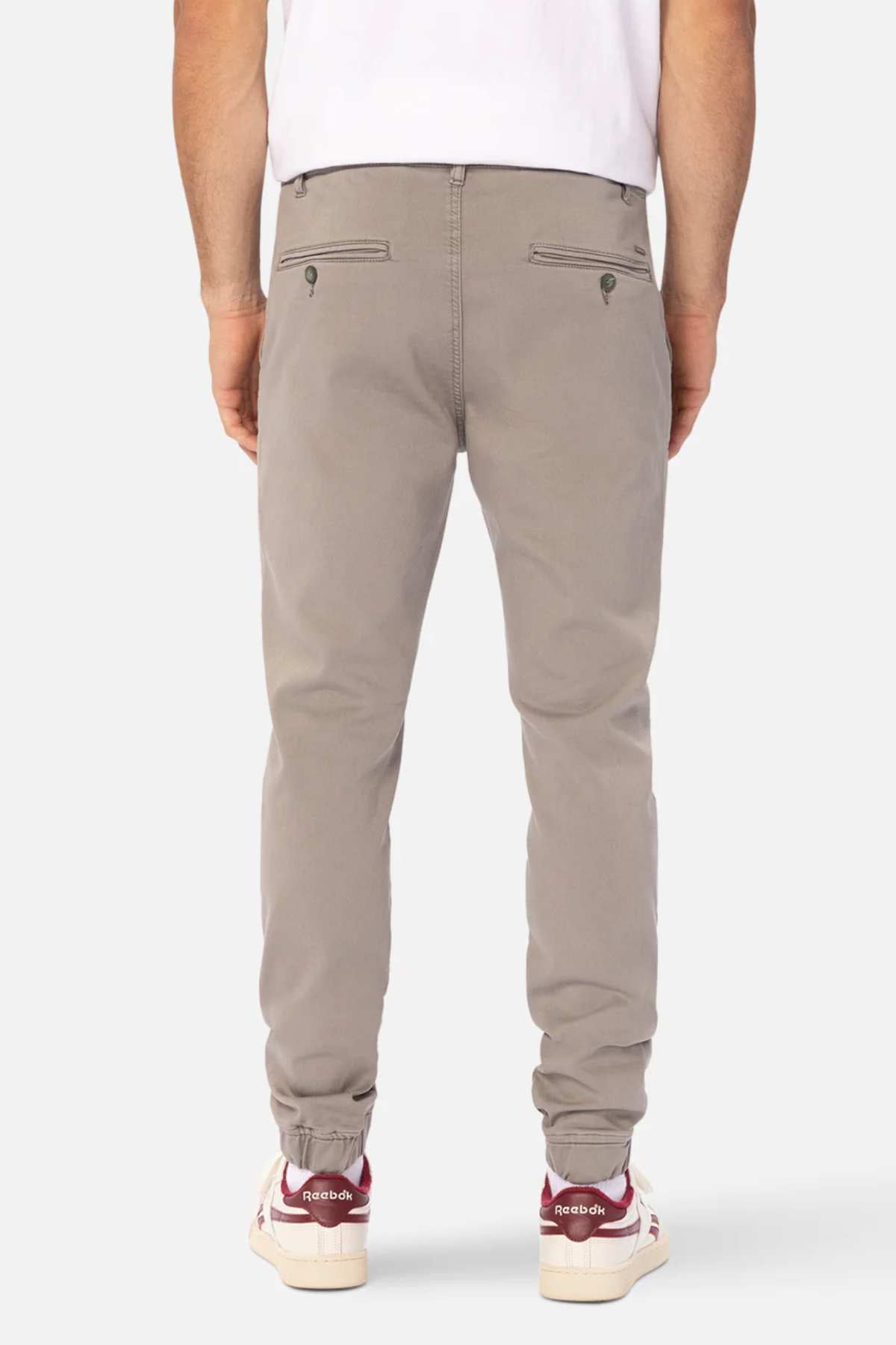 Someone is shown from the back wearing Industrie The Drifter Chino Pant in a slim fit light gray style with an elastic waist, paired with a white T-shirt and red-and-white Reebok sneakers.