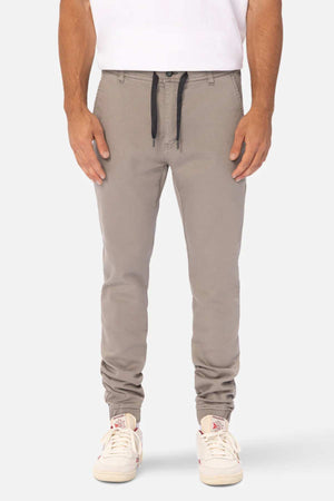 Someone wearing Industrie The Drifter Chino Pant in light gray with an elastic waist and black drawstring, paired with a white shirt and white sneakers, stands against a plain light background.