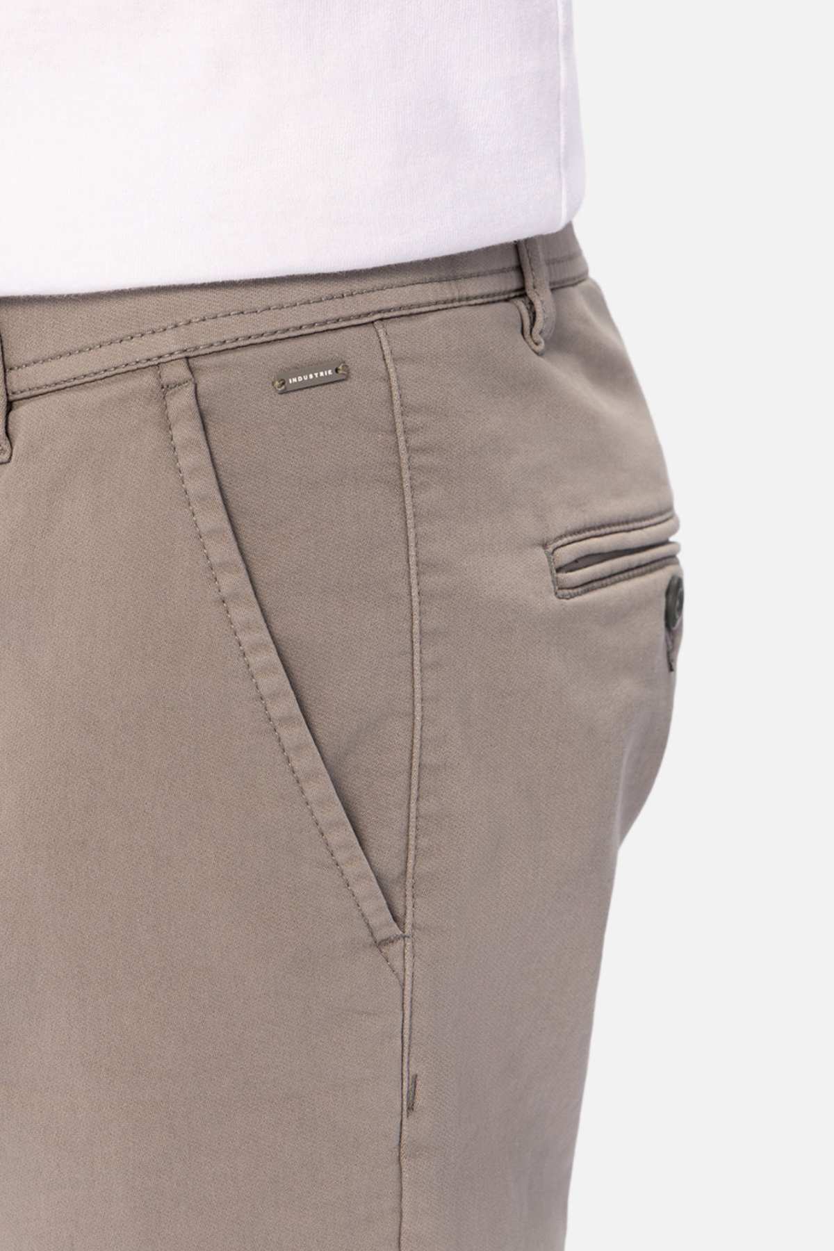Close-up of a person wearing Industrie The Drifter Chino Pant in slim fit light brown, styled with a tucked-in white shirt. Features include an elastic waist, side pockets, small logo tag, and a buttoned back pocket.