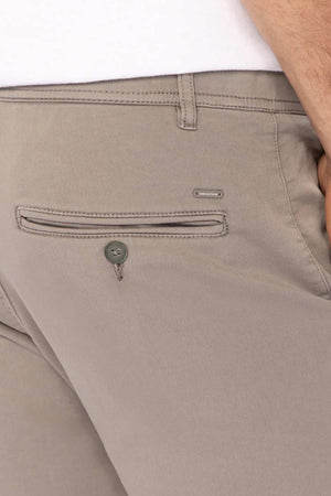 Close-up of a person wearing Industrie The Drifter Chino Pant in slim fit beige, highlighting the back pocket with button closure and a small metallic brand label above it. The person's hand rests near the pocket.
