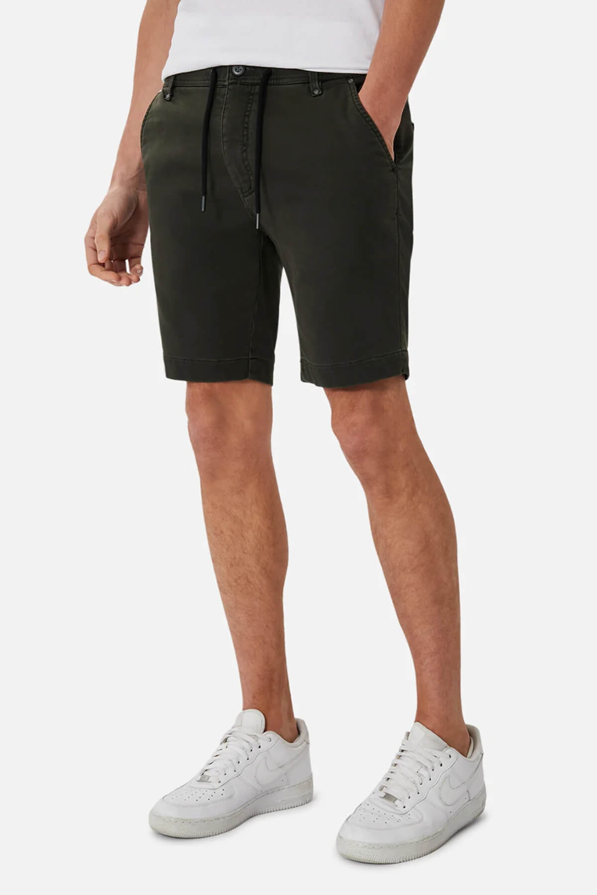 Sporting a stylish look, the person dons a white shirt with dark green Industrie The Drifter Cuba Shorts featuring a black drawstring, completing the ensemble with white sneakers. One hand casually in pocket, they exude effortless cool against the plain white backdrop.