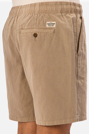 Wearing Industrie The Dublin Short in light brown, featuring an elastic waistband, a buttoned back pocket, and a small rectangular label above the pocket. Close-up showcases these summer shorts from the back and side.
