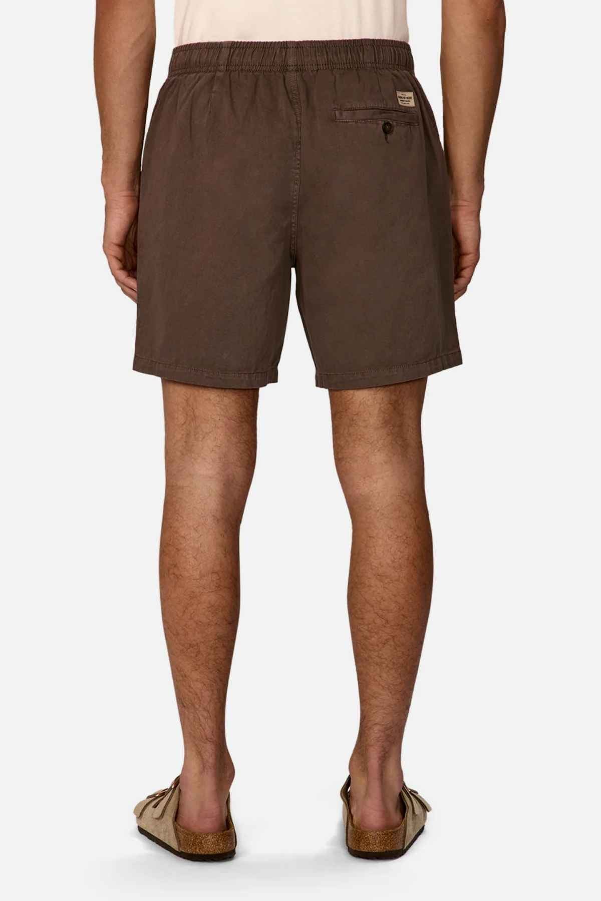 A person wearing the Industrie The Dublin Short, featuring a back pocket and button closure, stands with their back to the camera, highlighting their beige sandals. As a summer staple designed for comfort with an elasticised waistband, the shorts display the person's legs against a plain white background.