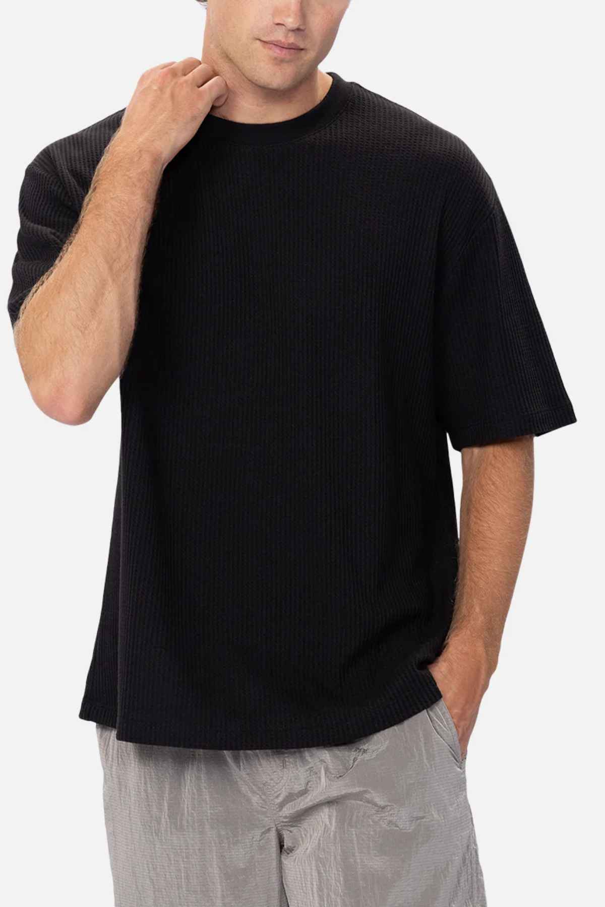 A person stands against a plain background wearing the Industrie The Earl Waffle Tee, a loose-fitting black t-shirt with a classic crew neck, paired with light gray pants. Their right hand is placed near their neck, and their left hand rests in their pants pocket. The overall mood is casual and relaxed.