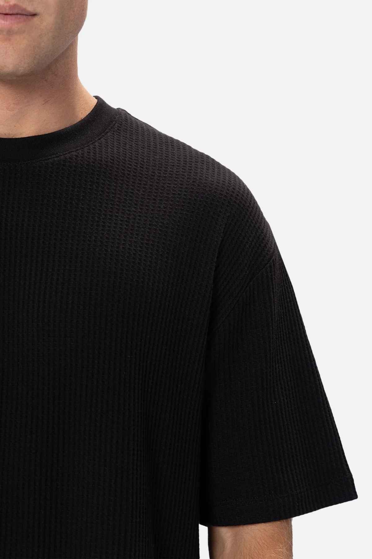 A close-up of a person wearing the Industrie The Earl Waffle Tee, a black, textured short-sleeve shirt featuring a ribbed pattern and a classic crew neck. Only the upper torso and part of the face are visible, set against a plain light background.