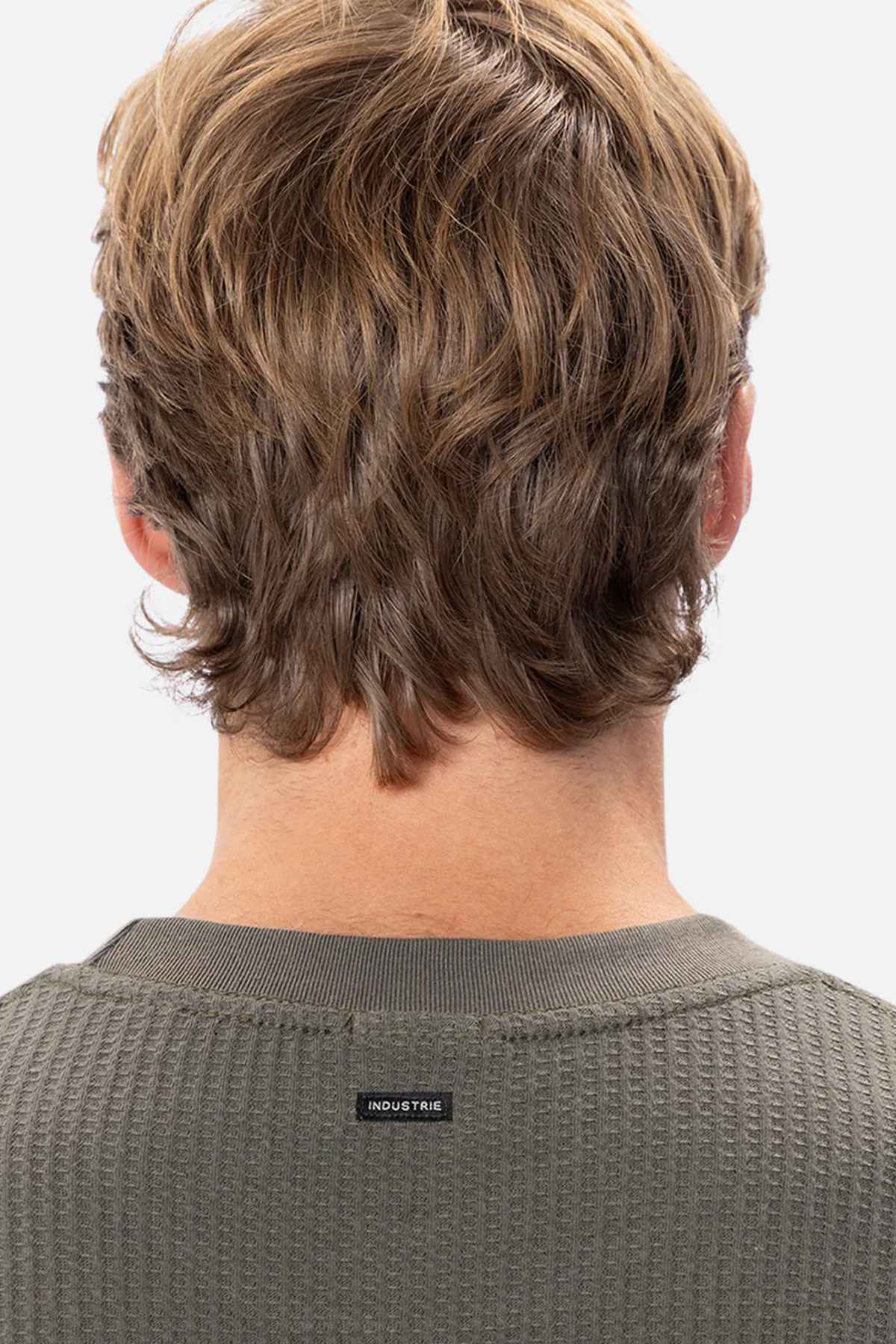 Back view of a person with wavy, light brown hair wearing a long-sleeved, green ribbed Industrie The Earl Waffle Tee. The focus is on the hair and the shirt's texture, with a small rectangular tag on the neckline reading "INDUSTRIE." The background is plain white.