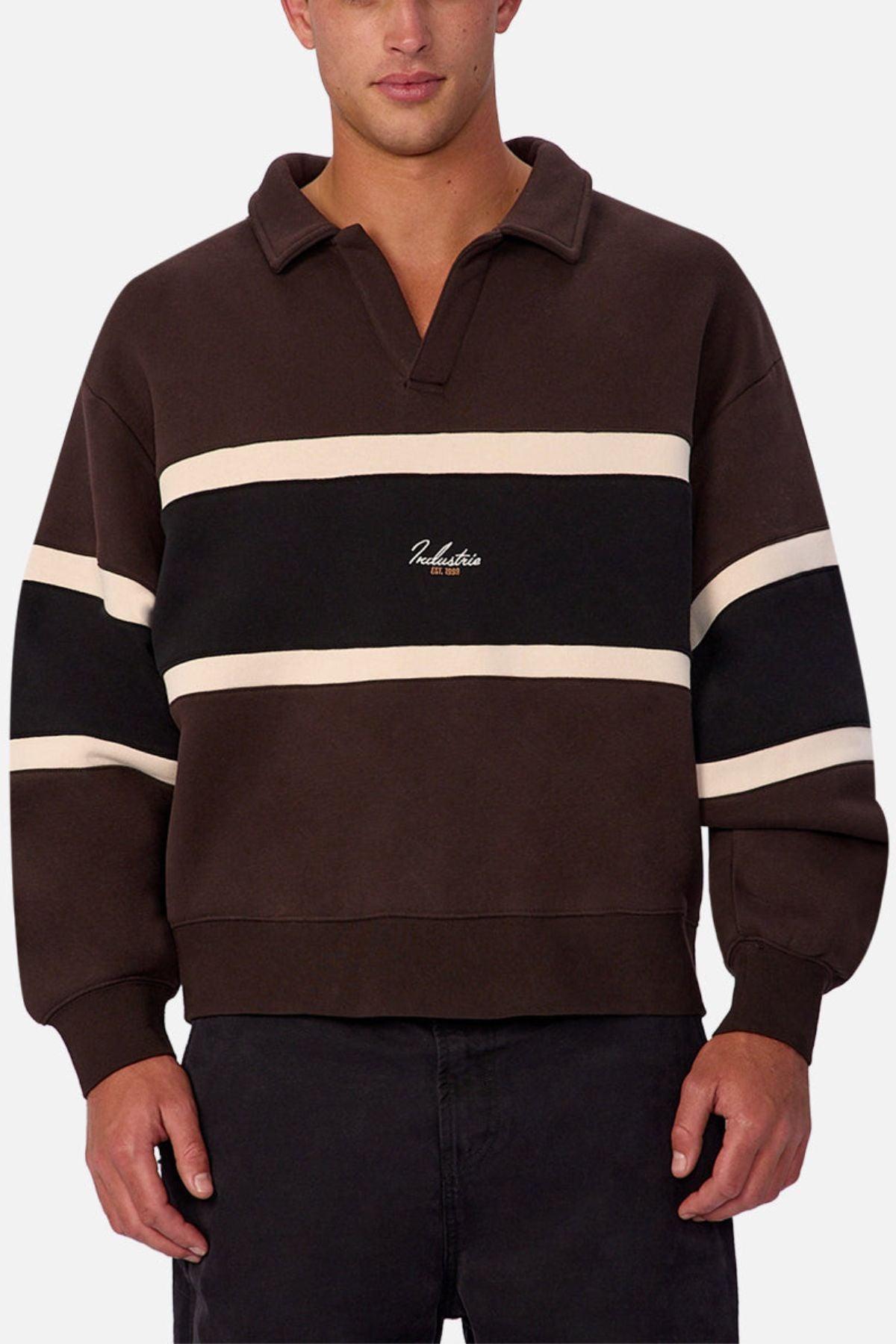 A man wears the Industrie The Eastbourne Tracktop—a dark brown, long-sleeve rugby-style shirt with cream and black stripes, collar, and white chest text—paired with dark pants for a relaxed, laid-back look.