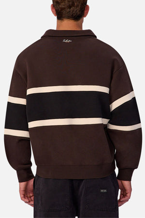 Someone is wearing the Industrie The Eastbourne Tracktop in dark brown, featuring black and white horizontal stripes on the back, paired with dark pants. The photo captures the relaxed silhouette’s back view against a light background.