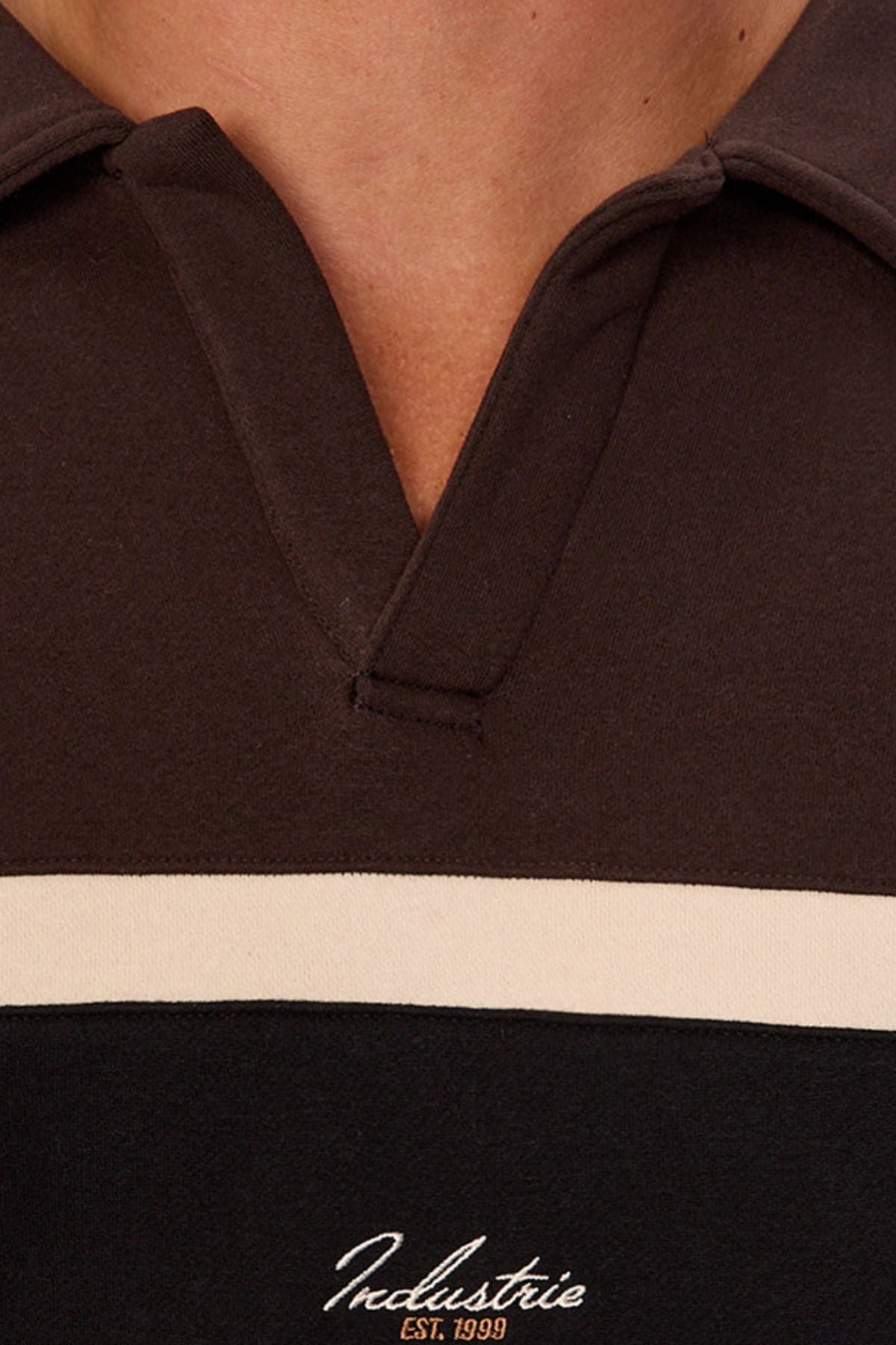 Close-up of a person's chest wearing the Industrie The Eastbourne Tracktop—a dark collared layer with a white horizontal stripe and "Industrie Est. 1999" embroidered below it, showcasing a relaxed silhouette.