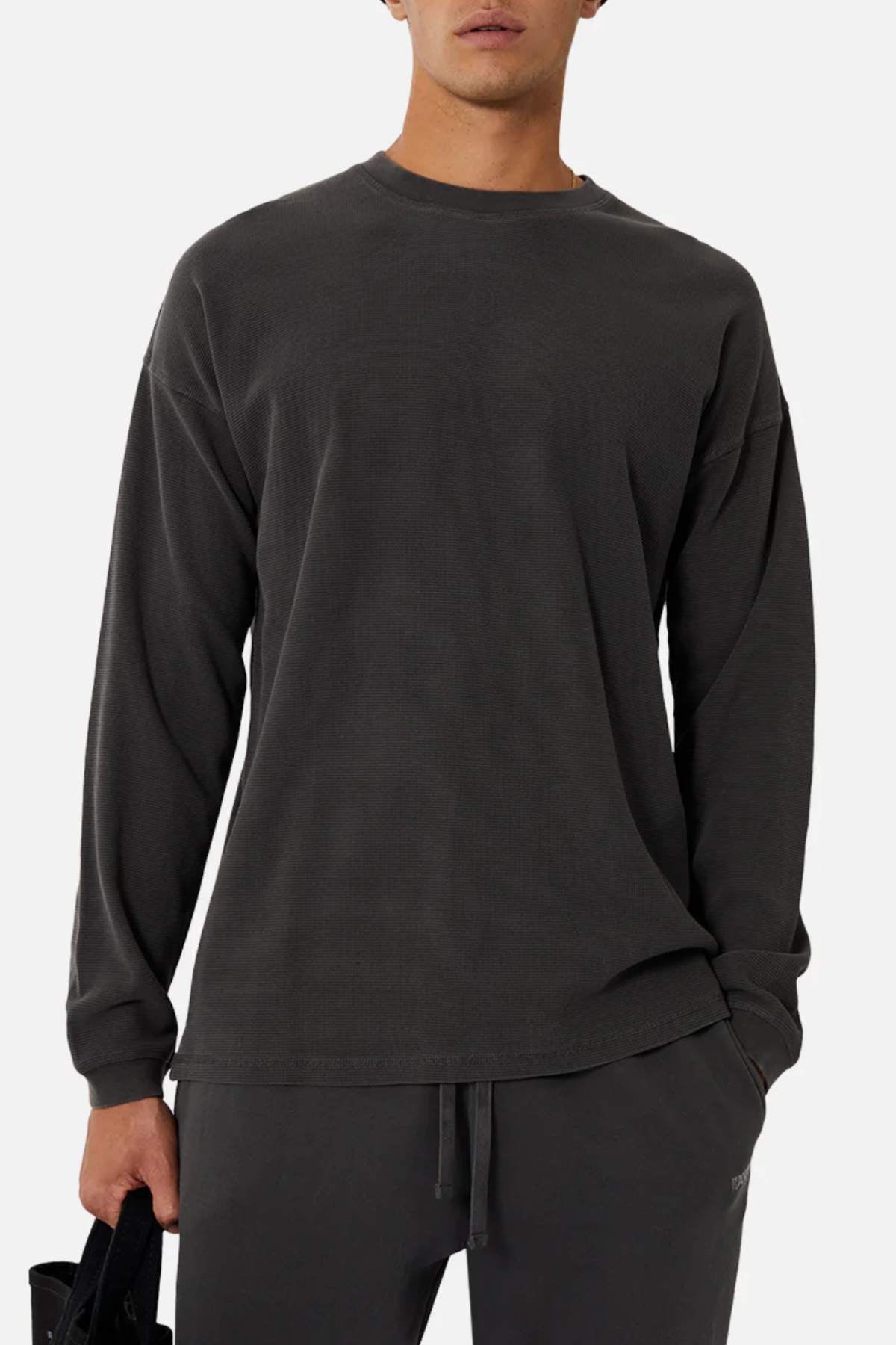 Wearing the Industrie The Eastport Waffle Long Sleeve Tee in dark gray with matching pants, someone stands against a plain background holding a small black bag in their left hand. Their head is slightly tilted, partially revealing their face.