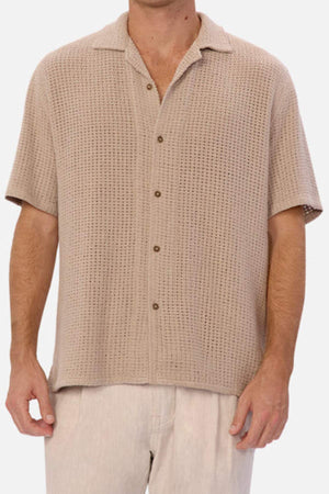 A man models the Industrie The Egnazia Short Sleeve Shirt, a beige, textured button-up made from a linen-cotton blend. Its breathable fabric ensures a relaxed fit for summer and pairs smoothly with light-colored pants against a plain white backdrop. Only his upper body and some pants are visible.