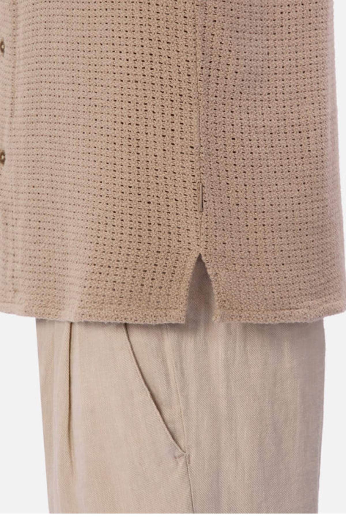 Close-up of a person wearing a beige textured cardigan with a split hem over light beige trousers. Highlighting the knit pattern of the linen-cotton blend cardigan and trouser pockets, these pieces are perfect additions to your breathable summer wardrobe alongside the Egnazia Short Sleeve Shirt.