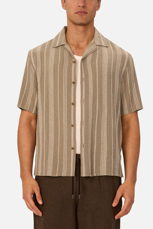 A person wearing the Industrie The Fabriano Short Sleeve Shirt—beige with vertical white stripes—over a white undershirt and brown pants, standing against a plain background.