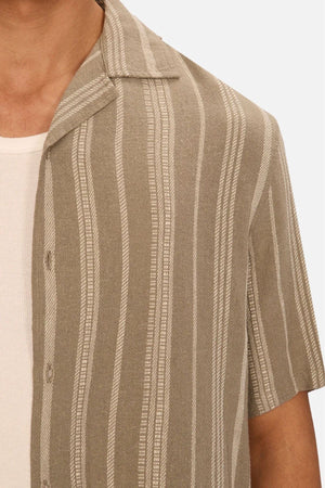 Close-up of a person wearing the Industrie The Fabriano Short Sleeve Shirt, a beige and white vertically striped lightweight cotton, camp-collar shirt over a white ribbed tank top. Only the upper torso and part of the left arm are visible.