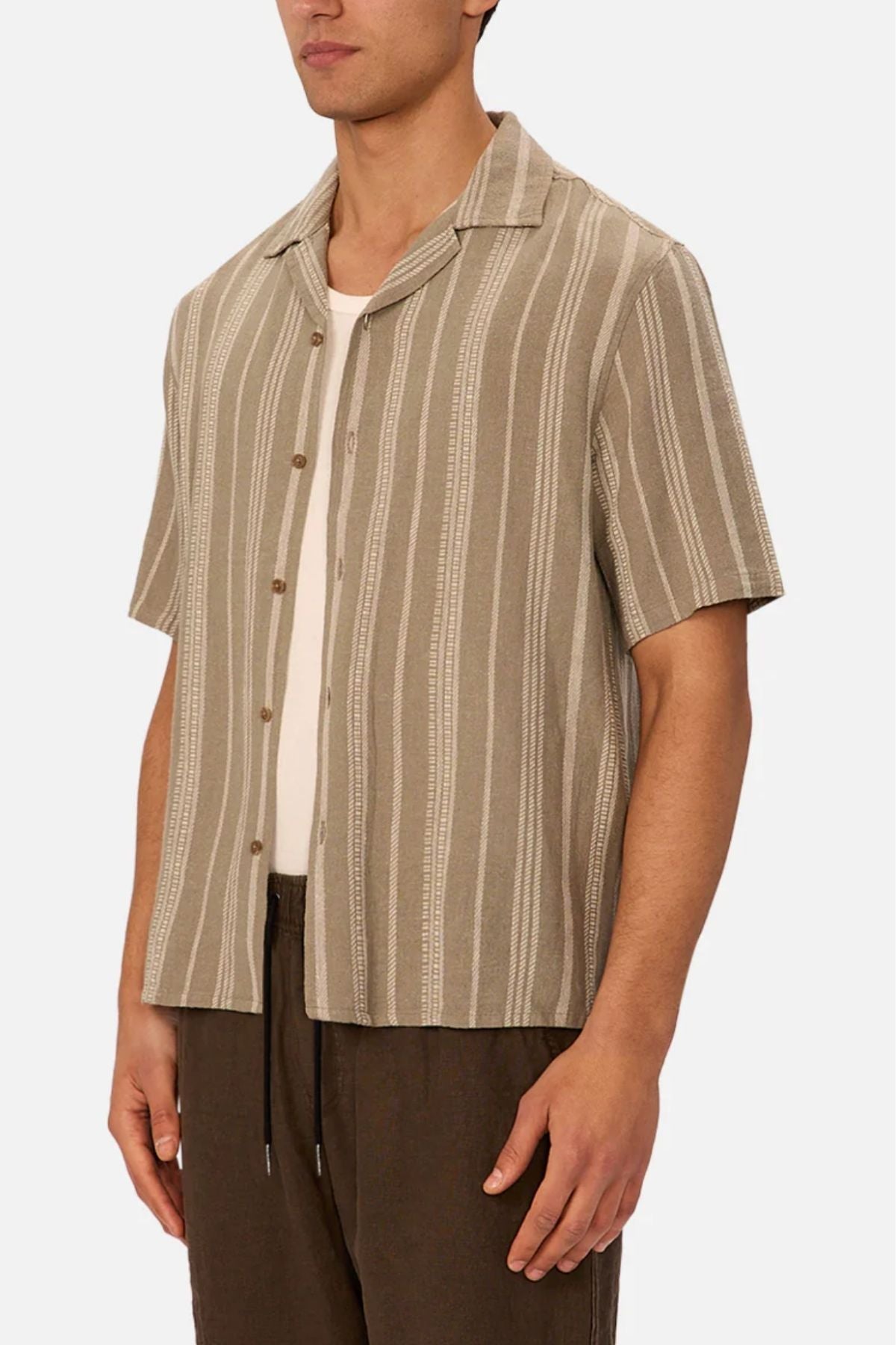 A man wears the Industrie The Fabriano Short Sleeve Shirt—a beige, relaxed fit camp collar shirt with vertical stripes—over a white T-shirt and dark brown pants against a plain background.