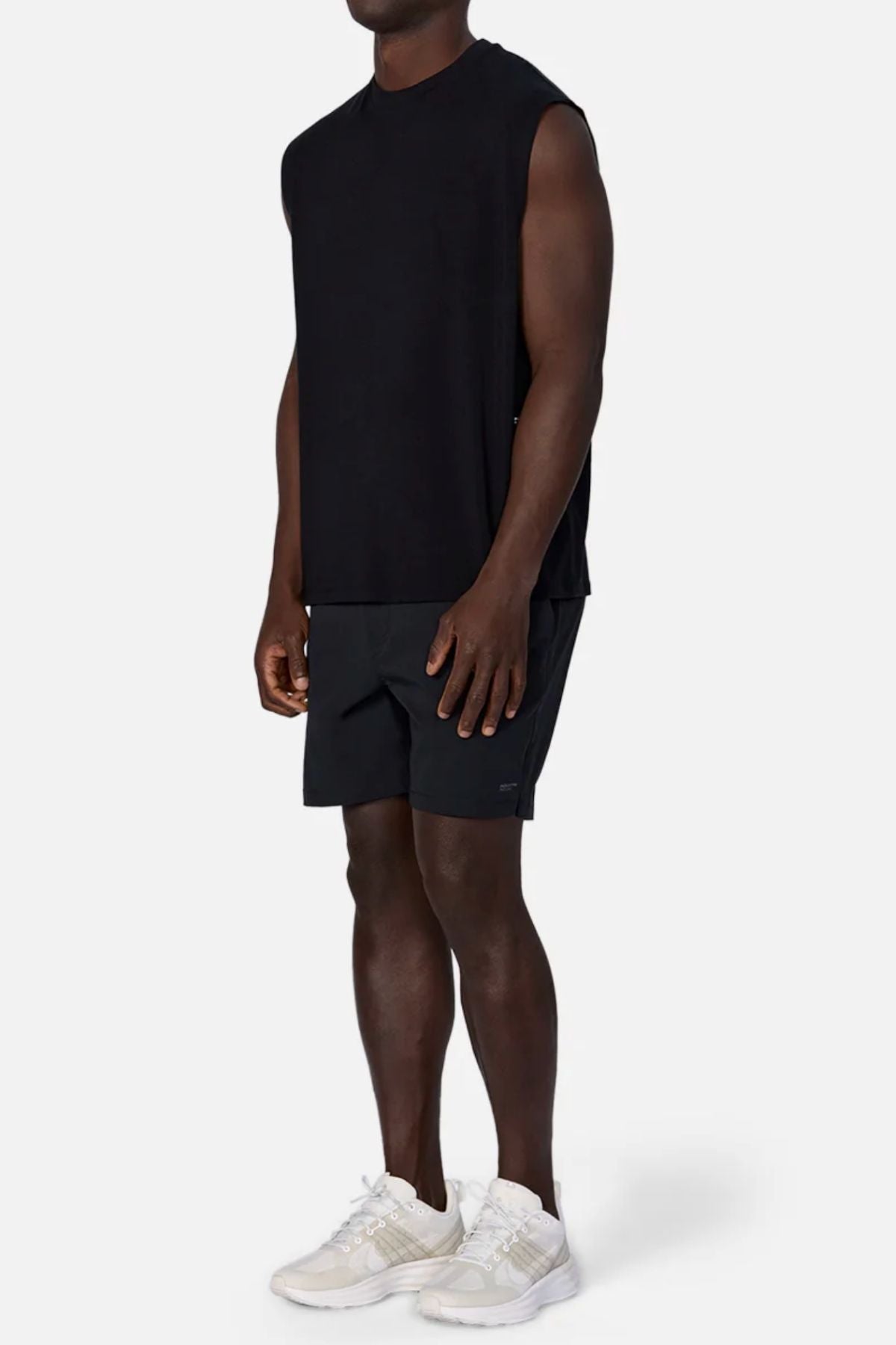 A man stands against a light background wearing the Industrie The Freeman Sleeveless Tee, black shorts, and white athletic shoes. His arms are relaxed at his sides and his face is not visible.