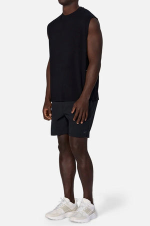 A man stands against a light background wearing the Industrie The Freeman Sleeveless Tee, black shorts, and white athletic shoes. His arms are relaxed at his sides and his face is not visible.