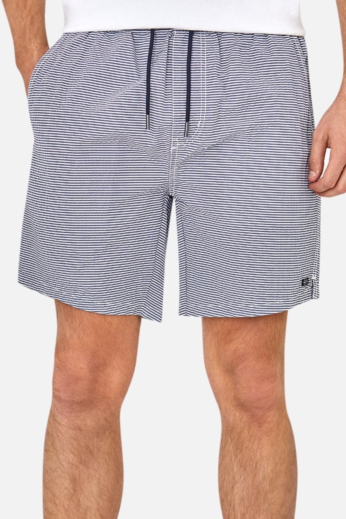 A person wears the Industrie The Freeport Bahama Short in white and navy blue striped seersucker, featuring an elasticised drawcord waist, pockets with hands inside, and a small left leg logo. Only part of the upper body is visible.