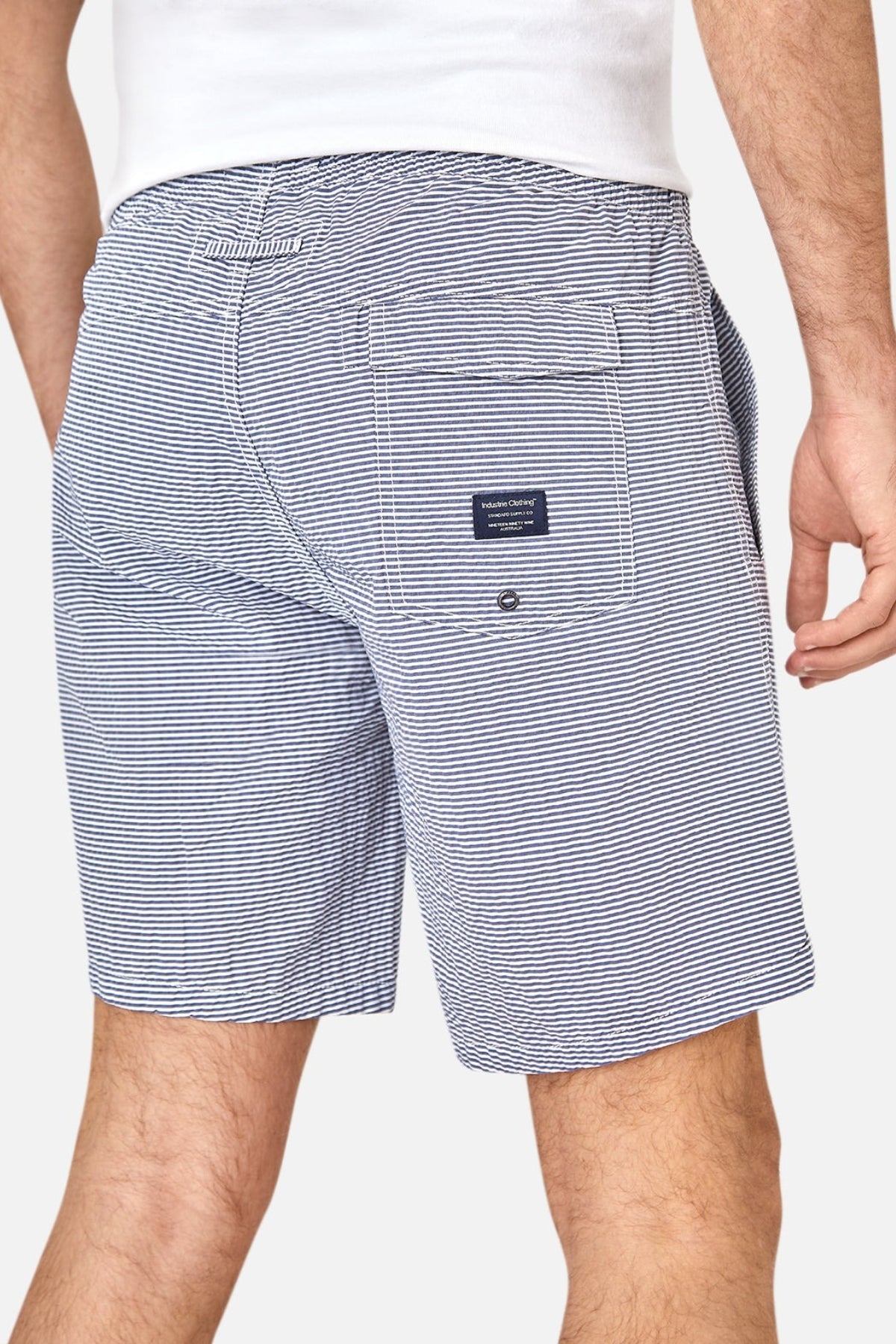 A man is shown from the waist down wearing Industrie The Freeport Bahama Short in blue and white seersucker with an elastic drawcord waist, a back pocket featuring a small navy label, and a white shirt; one hand relaxed by his side.