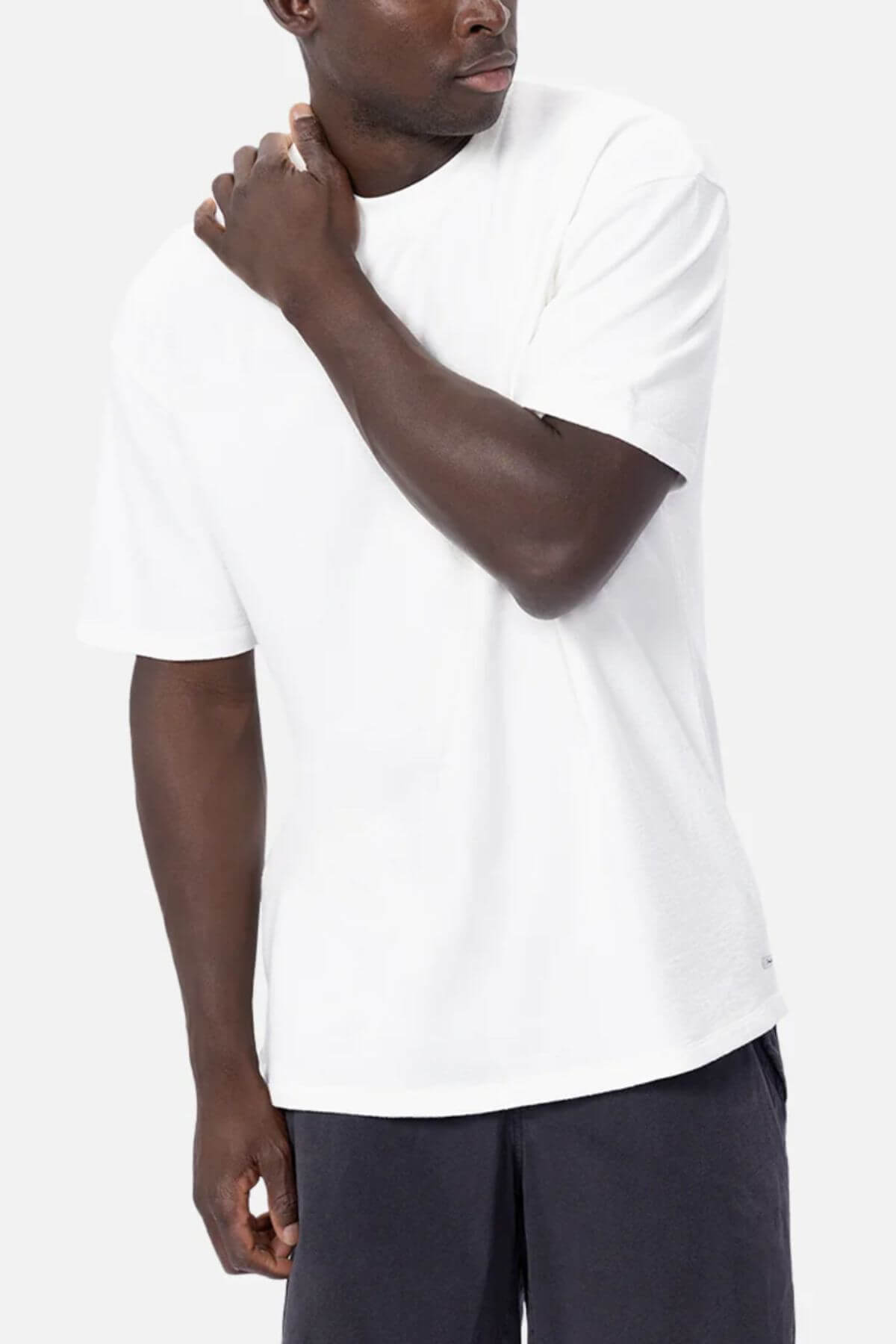 Dressed in an oversized fit, 100% cotton white Industrie The Henderson Boucle Tee and dark pants, a person stands against a light gray background, effortlessly draping the tee as they raise one hand to their neck and gaze aside.