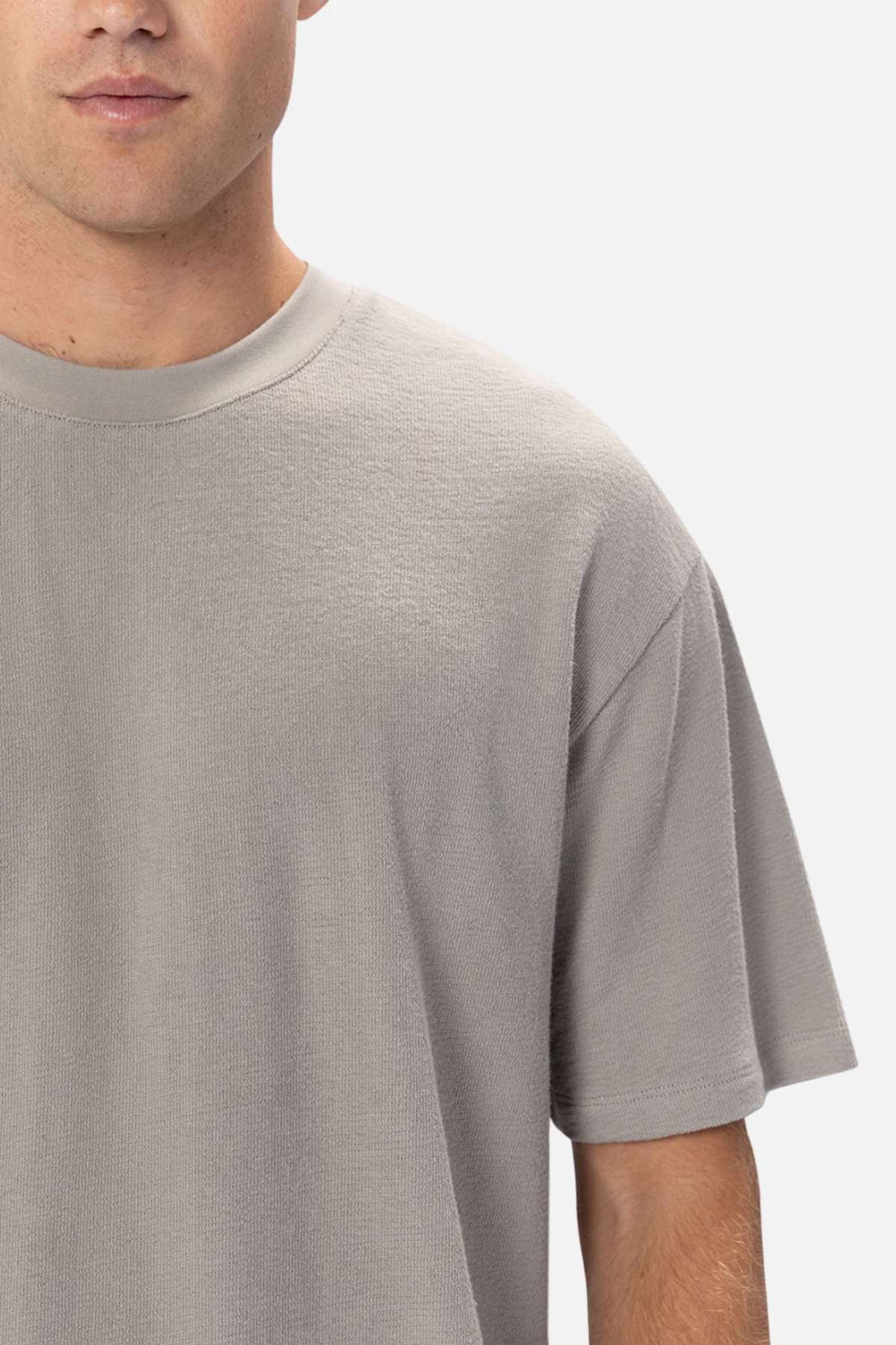 A partial view shows someone in an Industrie The Henderson Boucle Tee, featuring its oversized fit in plain gray 100% cotton. Their left face is partially hidden against a plain, light background.