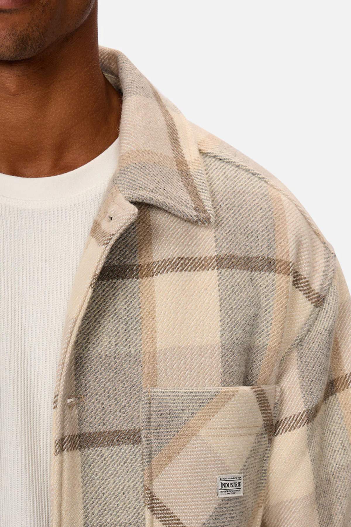 A person is wearing the Industrie The Hillside Jacket, displaying a plaid beige and grey check pattern over a white ribbed shirt. The jacket's top button is undone, revealing a "Industrie" label on the pocket. Focus is on the upper torso against a plain background.