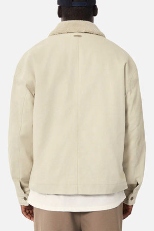 Seen from behind, a person wears Industrie The Huntsville Jacket in beige with a white collar over a white shirt, paired with a navy cap. The crisp backdrop accentuates the jacket's timeless appeal.