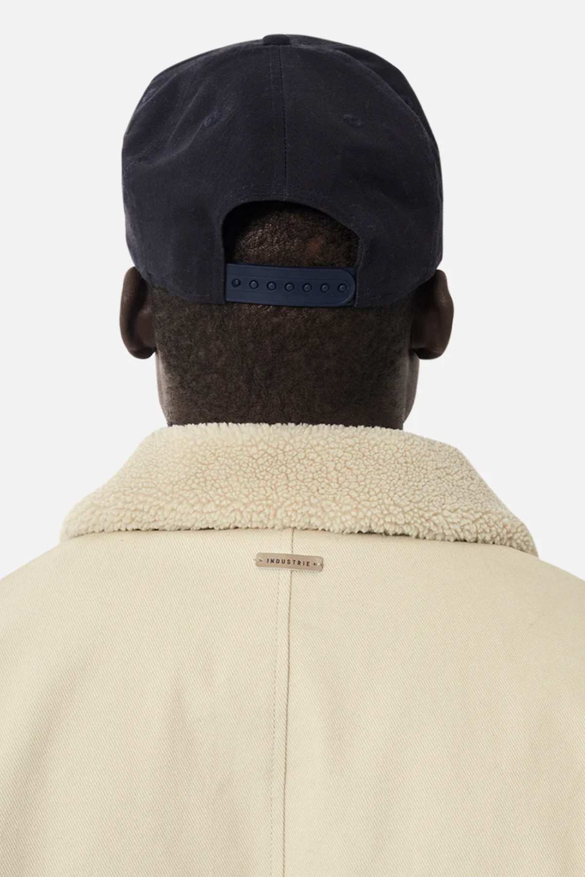 The image depicts the back of a person in a black baseball cap wearing the beige Industrie The Huntsville Jacket with sherpa lining and a classic collar against a plain white background.