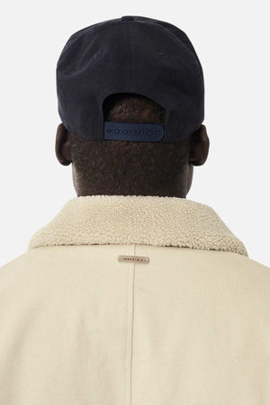 The image depicts the back of a person in a black baseball cap wearing the beige Industrie The Huntsville Jacket with sherpa lining and a classic collar against a plain white background.