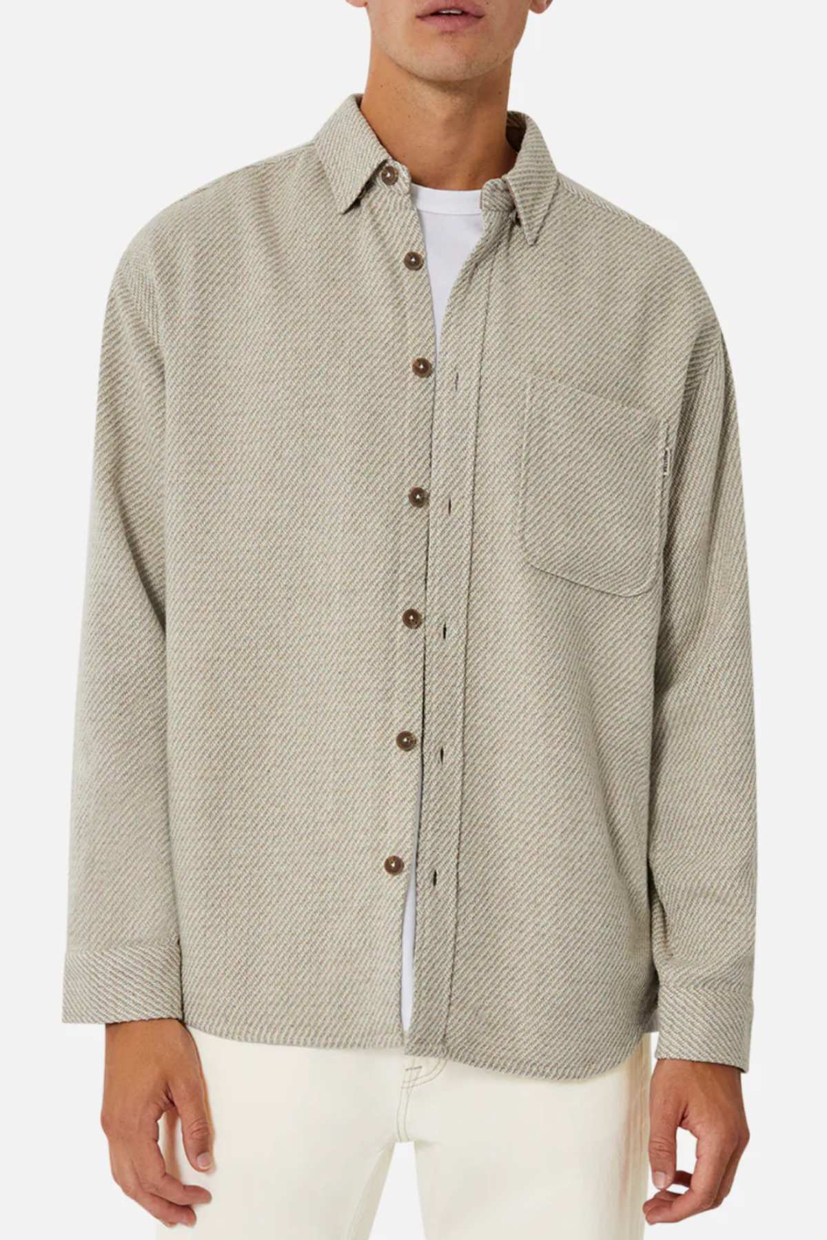 A man wears the Industrie The Jackson Shirt in light gray with a subtle herringbone pattern, featuring a chest pocket and brown buttons, over a white T-shirt. This stylish long-sleeved shirt is paired with light-colored pants for a relaxed fit.
