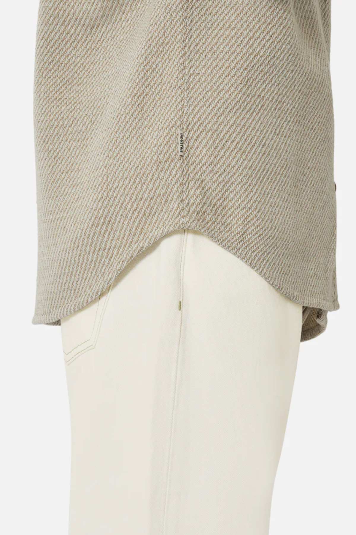 A close-up shows a person in the Industrie The Jackson Shirt, a light beige knit top with a relaxed fit over white pants. It boasts a textured, slightly curved hem and a small tag on the edge, set against a plain white background.