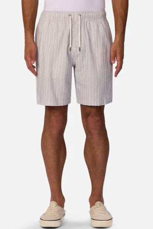 A person wearing Industrie The LA Brava Short—light gray and white vertical striped linen blend men’s casual shorts with a drawstring waist—paired with a white shirt and slip-on shoes, stands against a plain white background.