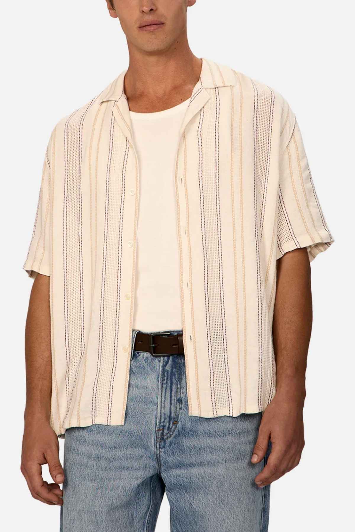 A person wearing the Industrie The Lakeville Short Sleeve Shirt, which features a cream and beige textured stripe design, over a white T-shirt, paired with light blue jeans and a black belt, stands against a neutral background.