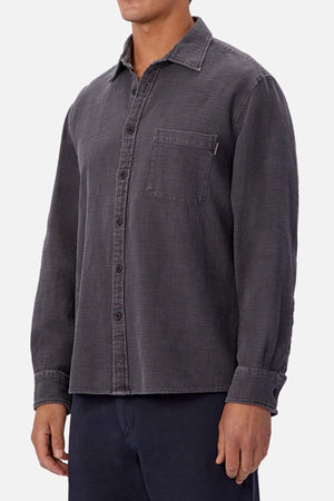 A man wears the Industrie Lisbon Long Sleeve Shirt in dark gray, featuring a classic collar and chest pocket, paired with dark pants. He is shown from the thighs up against a plain white background.