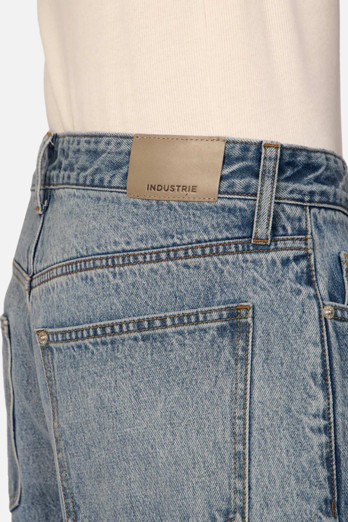 Close-up of a person wearing Industrie The Lone Star Jean in light blue denim, featuring a leather patch on the back waistband labeled "INDUSTRIE." The person is also wearing a light-colored shirt tucked into the jeans. The focus is on the back pocket and the brand label.
