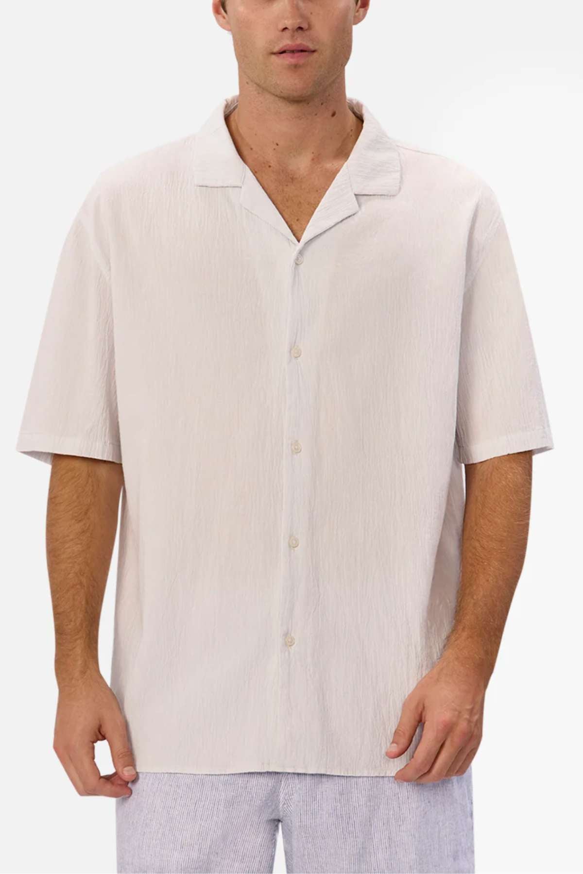 A man models the Industrie The Magna Short Sleeve Shirt, a light beige summer top crafted from 100% cotton. It features a relaxed collar and subtle texture with an oversized fit, set against a blank backdrop and paired seamlessly with light-colored pants.