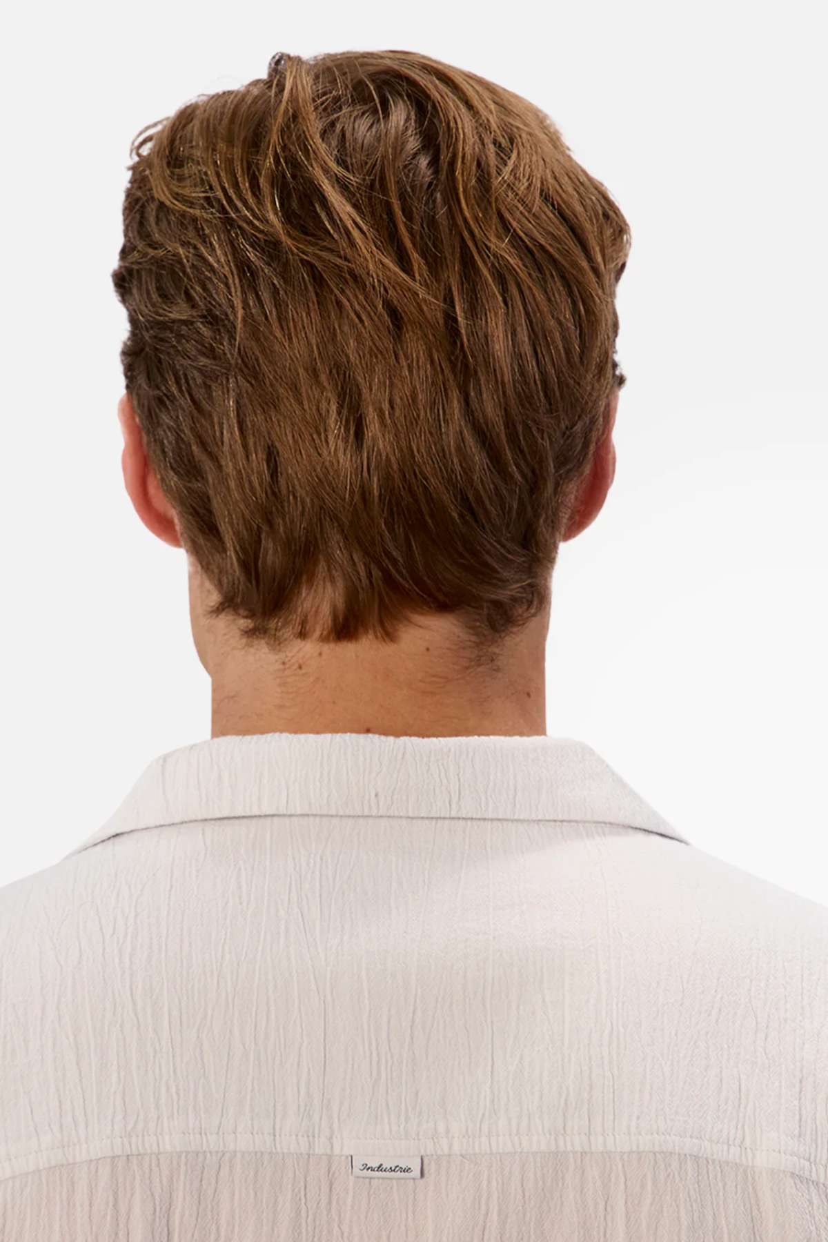 The image shows the back of a person's head and shoulders with short light brown hair, wearing the oversized fit Industrie The Magna Short Sleeve Shirt in white. A small tag is near the collar, and the plain white background highlights the 100% cotton fabric's crisp elegance.