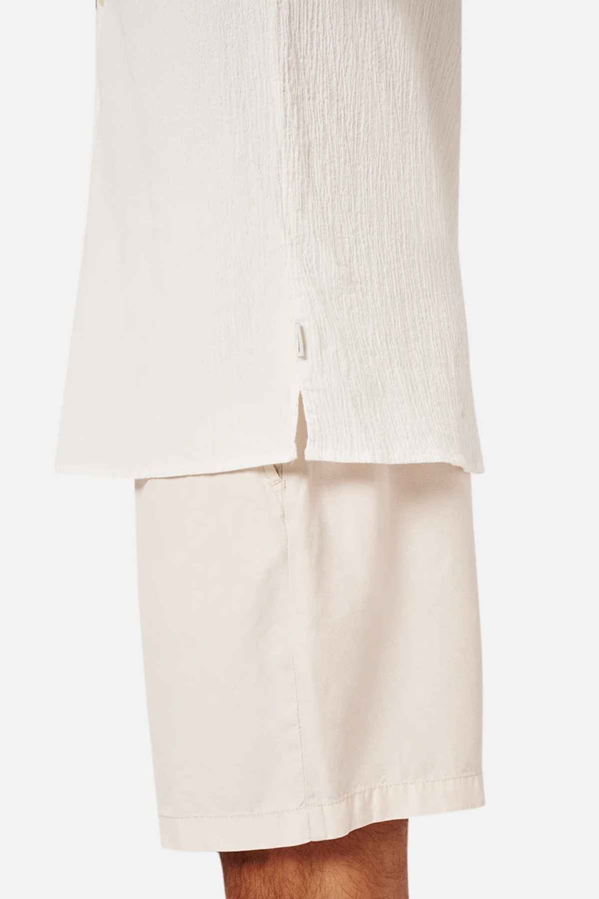 A close-up view of a person wearing the Industrie The Magna Short Sleeve Shirt in light beige with a side slit and matching light beige shorts. The breathable cotton fabric highlights part of the arm and leg, making it perfect for warm weather. The background is plain white.