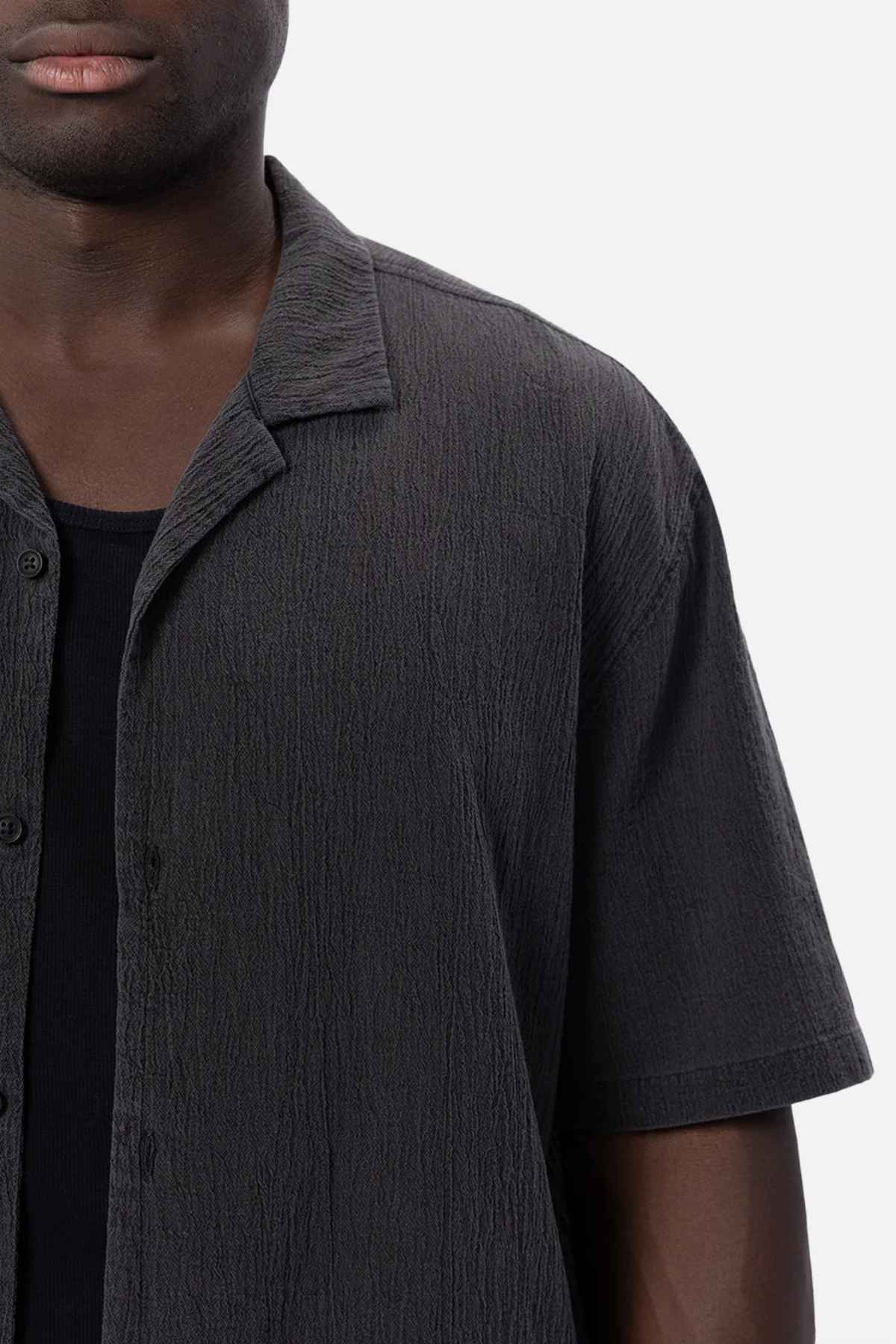 A person with dark skin is shown wearing the Industrie The Magna Short Sleeve Shirt, a textured black shirt featuring an oversized fit and short sleeves. It is layered over a black top, crafted from what appears to be 100% cotton, against a plain white background.