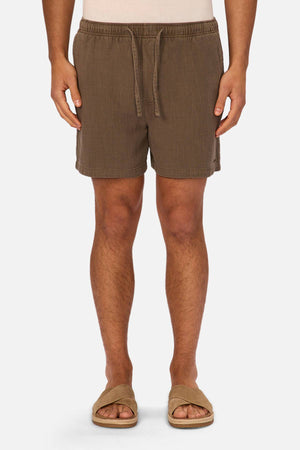 A person is seen from the waist down wearing Industrie The Mallorca Short, made of lightweight textured fabric with an elasticated waist, paired with tan slide sandals, standing against a plain white background.